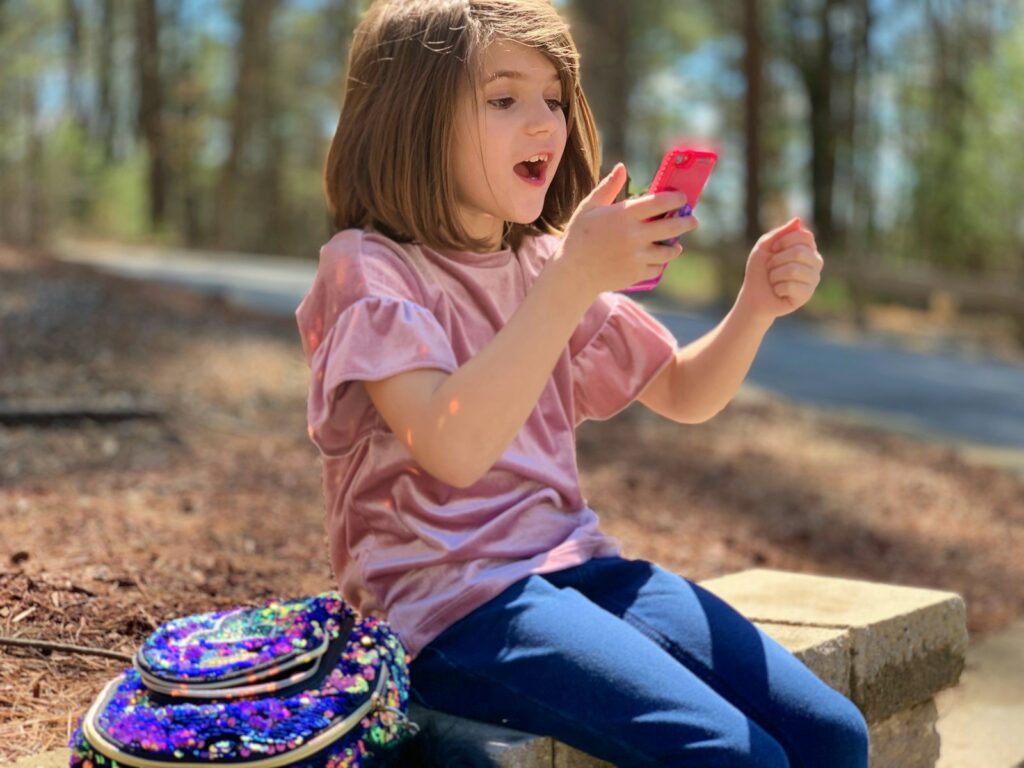 A young girl sits on a stone bench outdoors, smiling in surprise at the pink phone in her hands