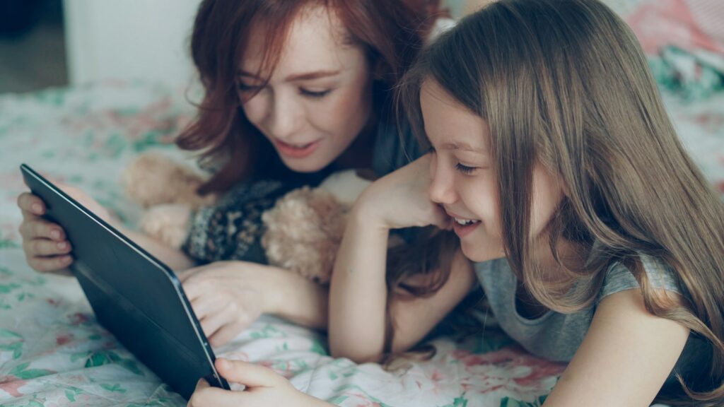 A mother and her young daughter lie on a bed and smile as they watch a tablet together