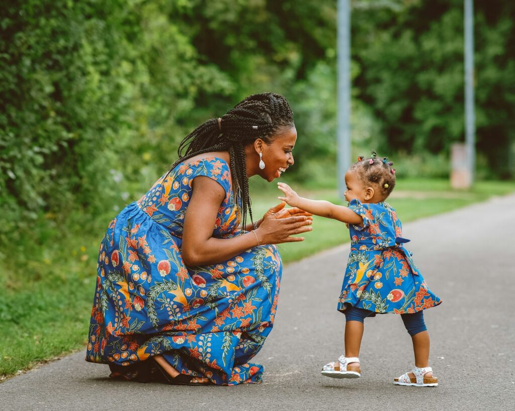 A smiling mother in a bright patterned dress kneels to meet her toddler daughter on a park path