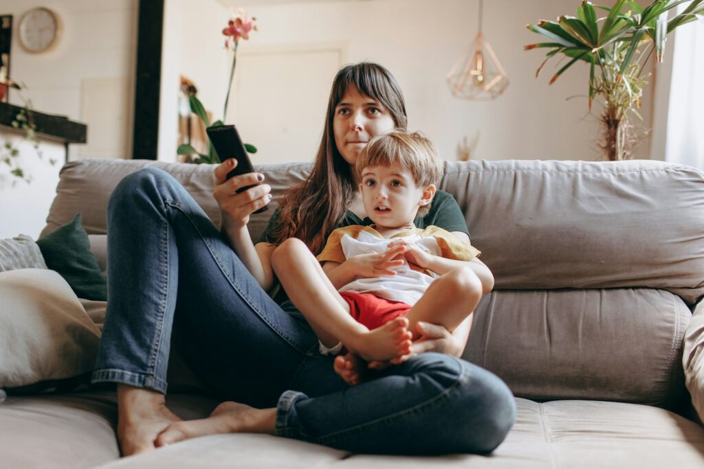 A mother sits on a couch with her toddler on her lap, holding her phone