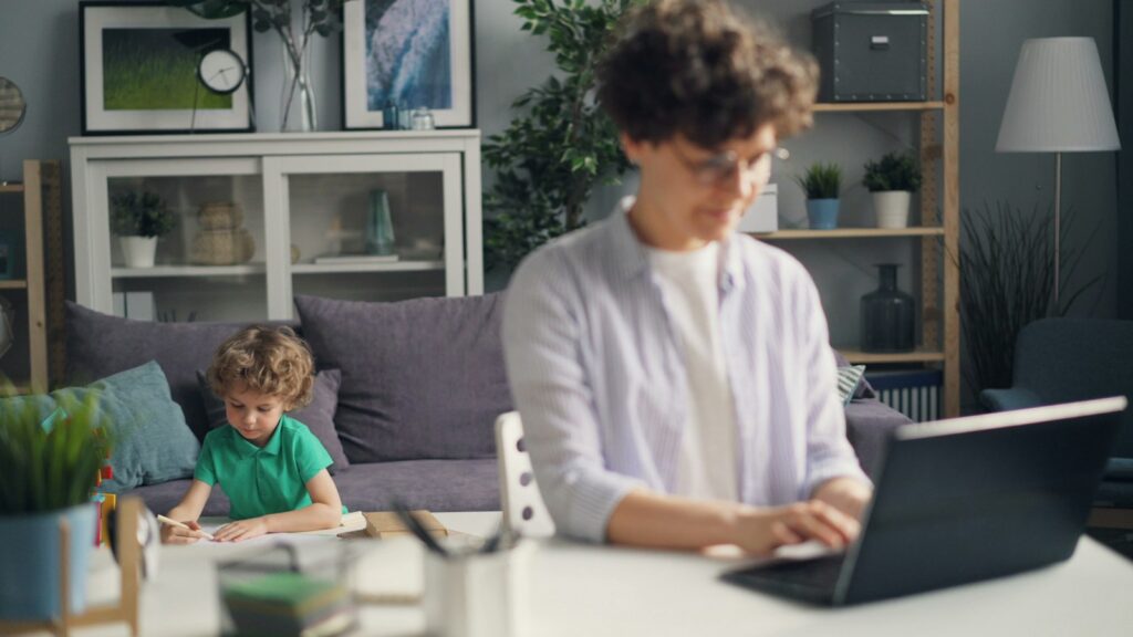 A woman works on a laptop at a table at home while her young son draws on the sofa behind her