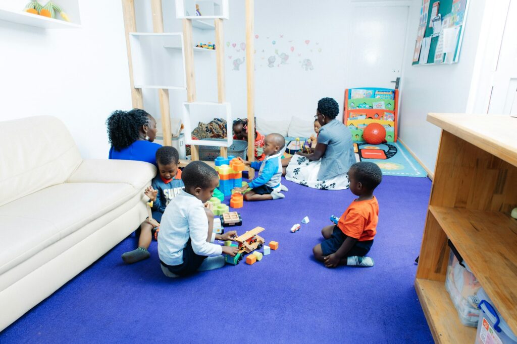 Young children and two adults play with blocks and toys on a blue carpet in a bright childcare room