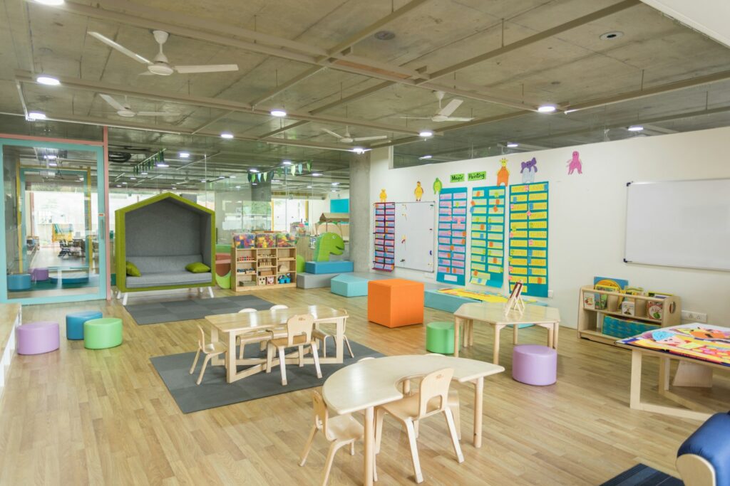 A bright, open childcare room with small tables and chairs, a reading nook and colorful floor seats