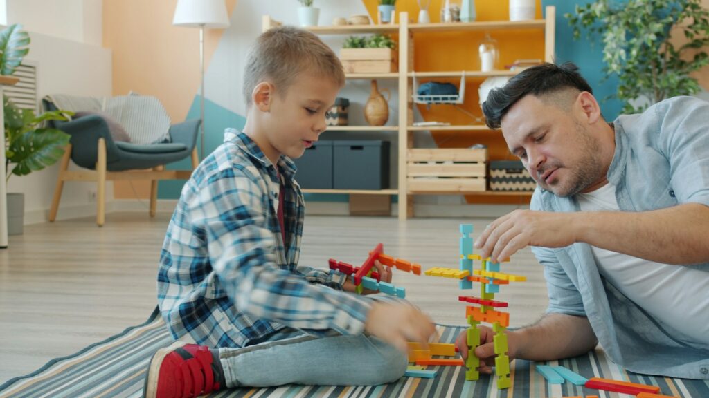A father and his young son building a tower of colorful blocks together on the living room floor.