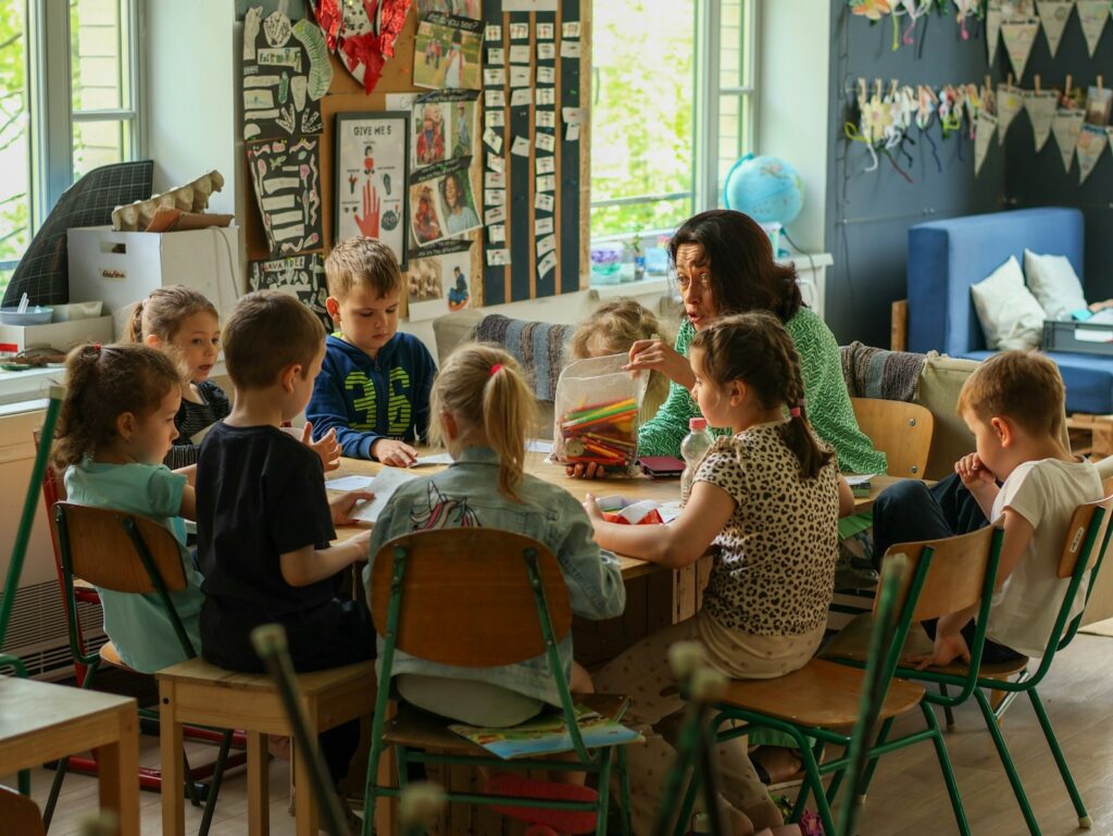 A teacher sits with a group of young children around a table in a bright, cozy classroom