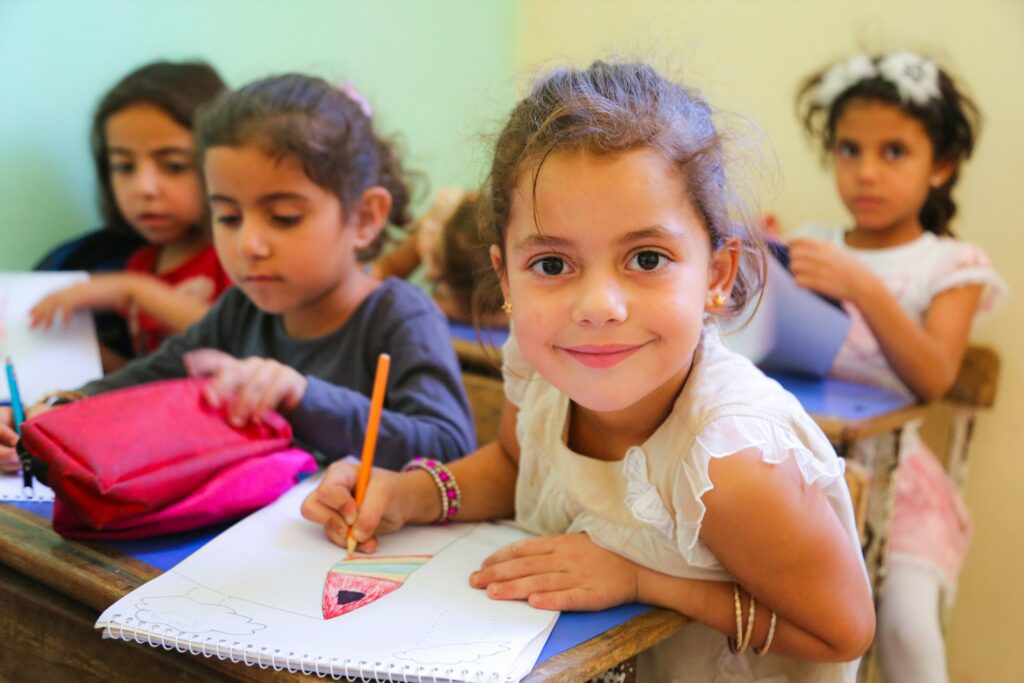 A smiling girl draws at her school desk while other young children work behind her