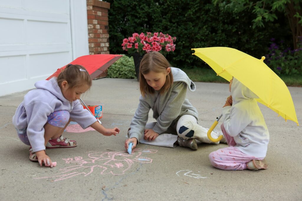 Children crouch together drawing with chalk on a driveway under bright umbrellas.