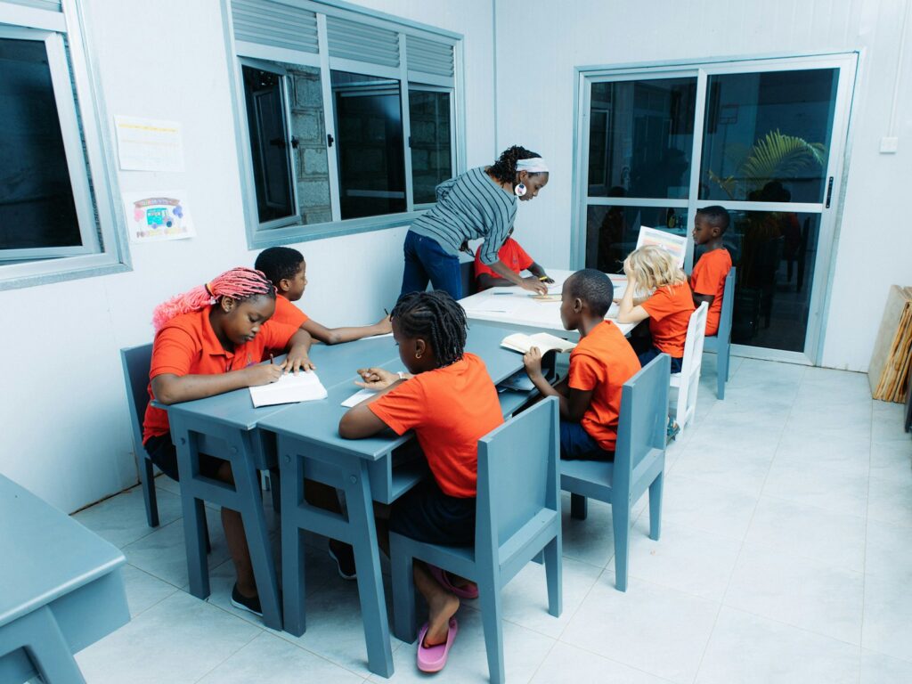 A teacher leans over a table to help a group of children with their work in a bright classroom