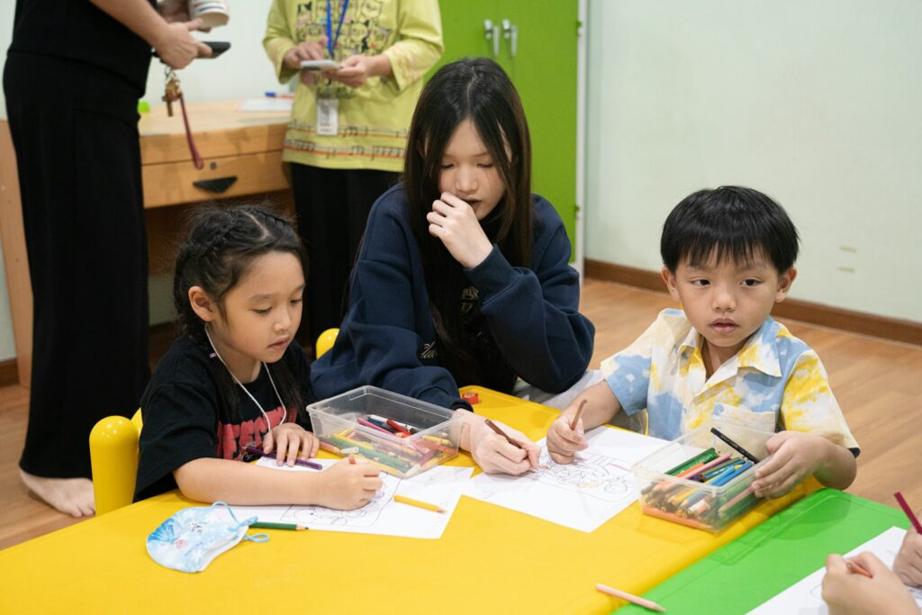 A teacher helps three children draw with colored pencils at a bright yellow classroom table