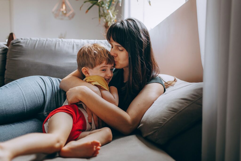 A mother cuddles her toddler on the couch and kisses the top of his head