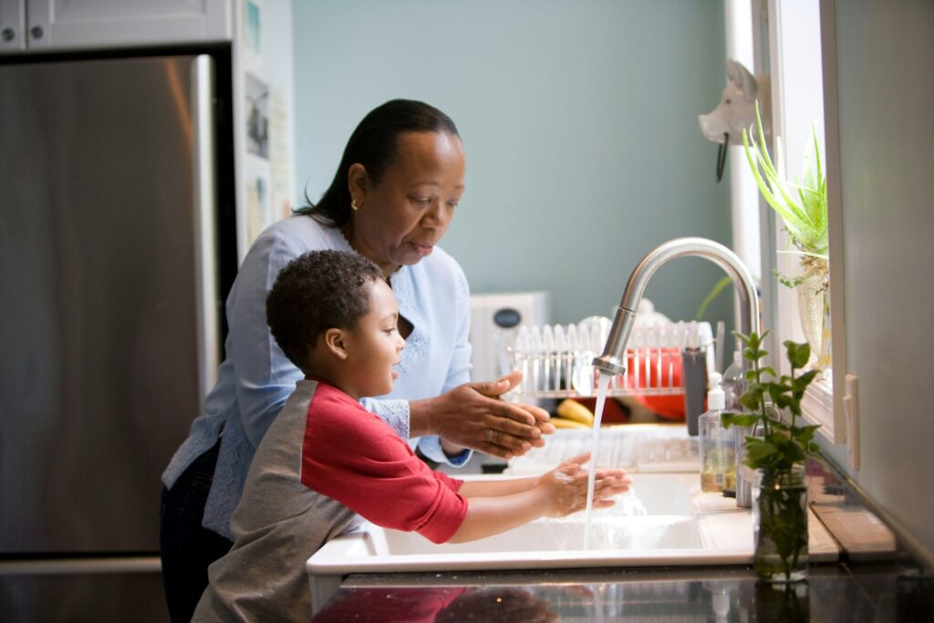 A woman helps a young boy wash his hands at a kitchen sink