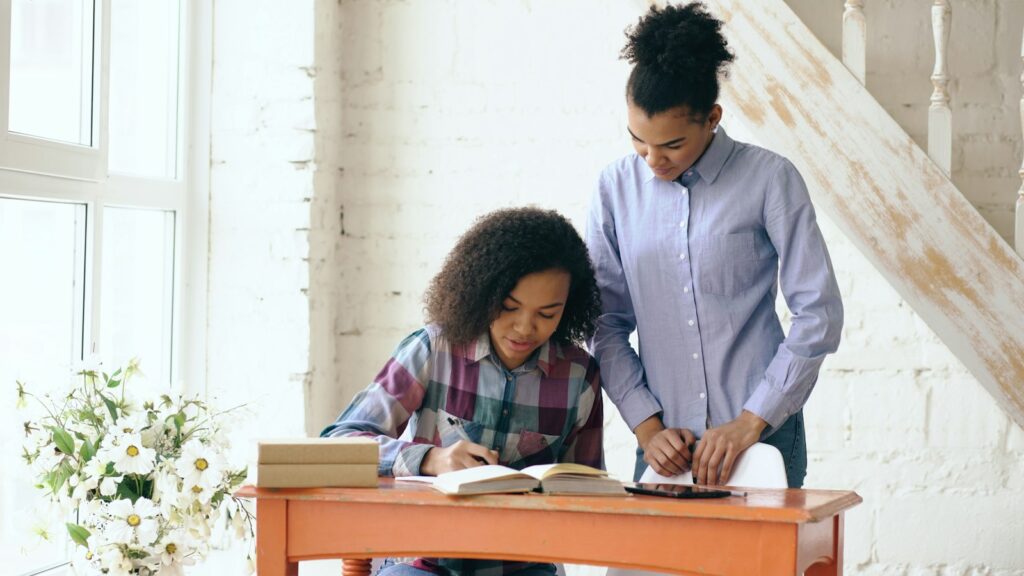 A woman stands beside a girl and helps her with homework at a wooden desk