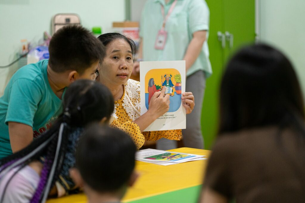 A teacher holds up an illustrated picture book for a small group of children at a table