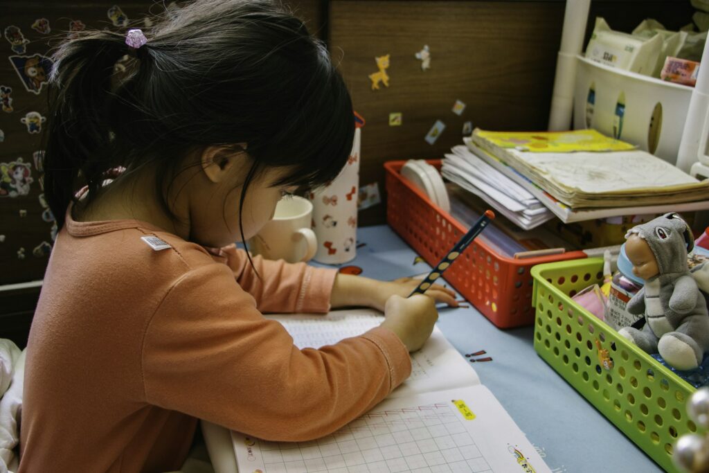 A young girl in an orange top concentrates on writing in a notebook at her desk, with books and a stuffed toy nearby