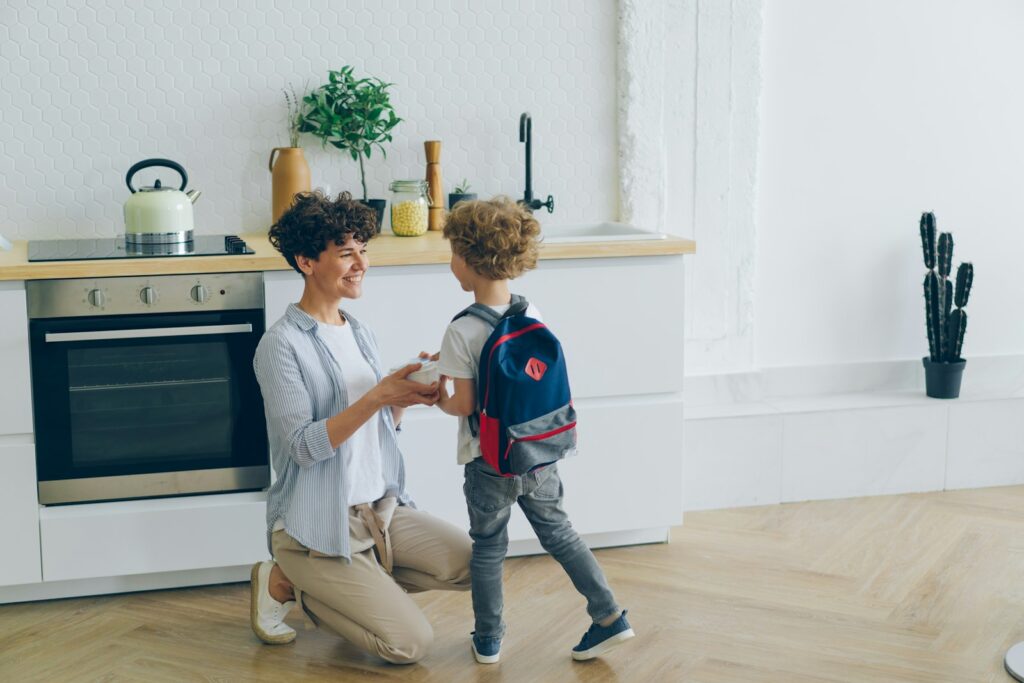 A smiling mother kneels in the kitchen to hand her young son a lunch box before he heads out with his backpack