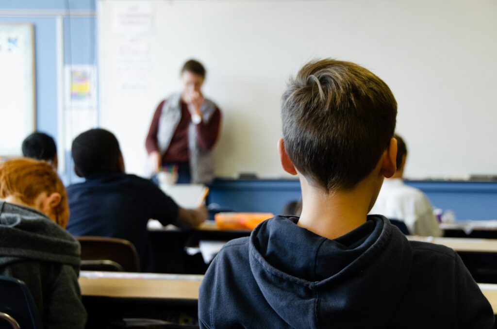 A student in a dark hoodie, seen from behind, faces a teacher at the whiteboard in a school classroom