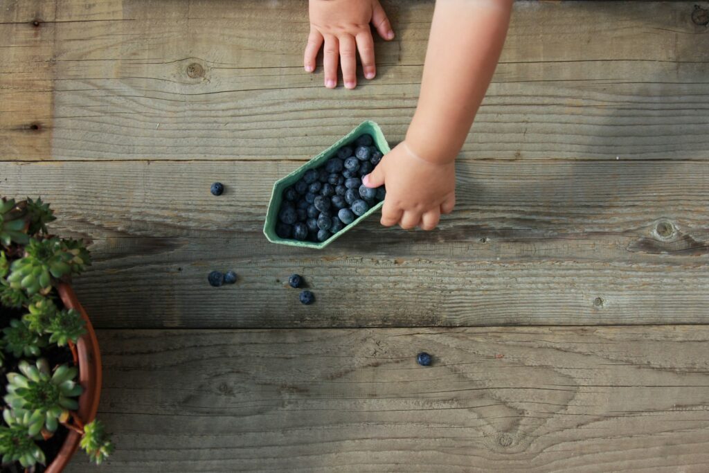 A toddler's hand reaches into a small basket of fresh blueberries on a rustic wooden table
