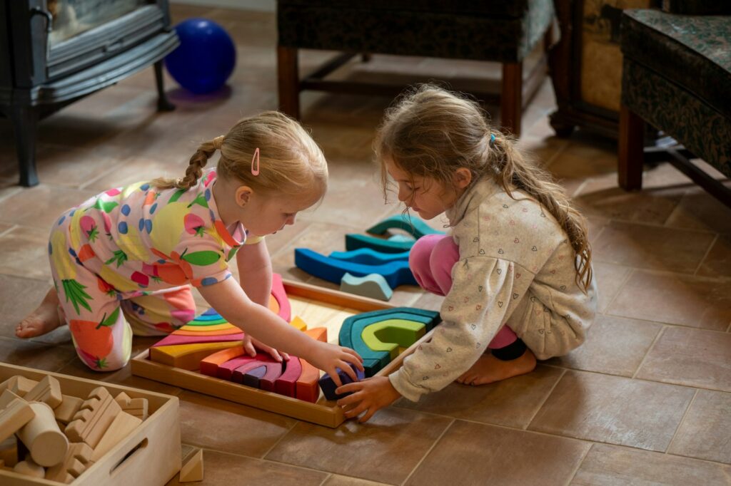 Two young girls kneel on the floor and play with colorful wooden shapes together