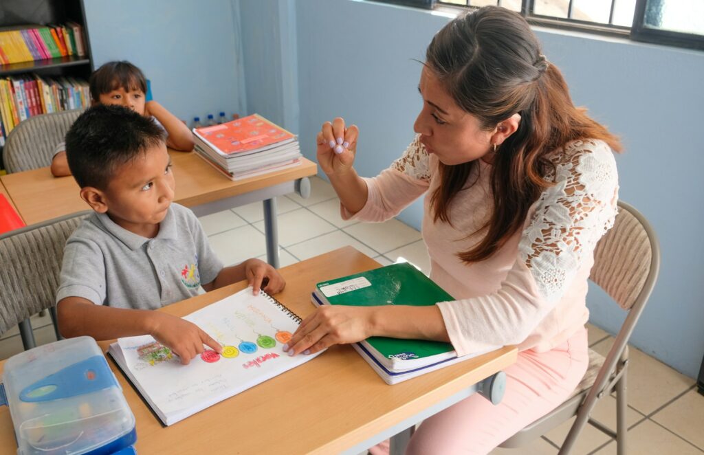 A teacher sits at a table with a young boy and gestures as they look at his workbook together