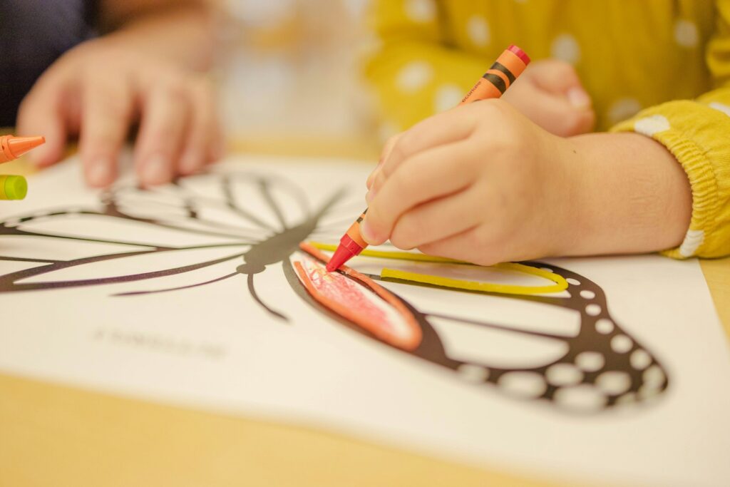 A child's hand coloring a butterfly outline with an orange crayon at a classroom table