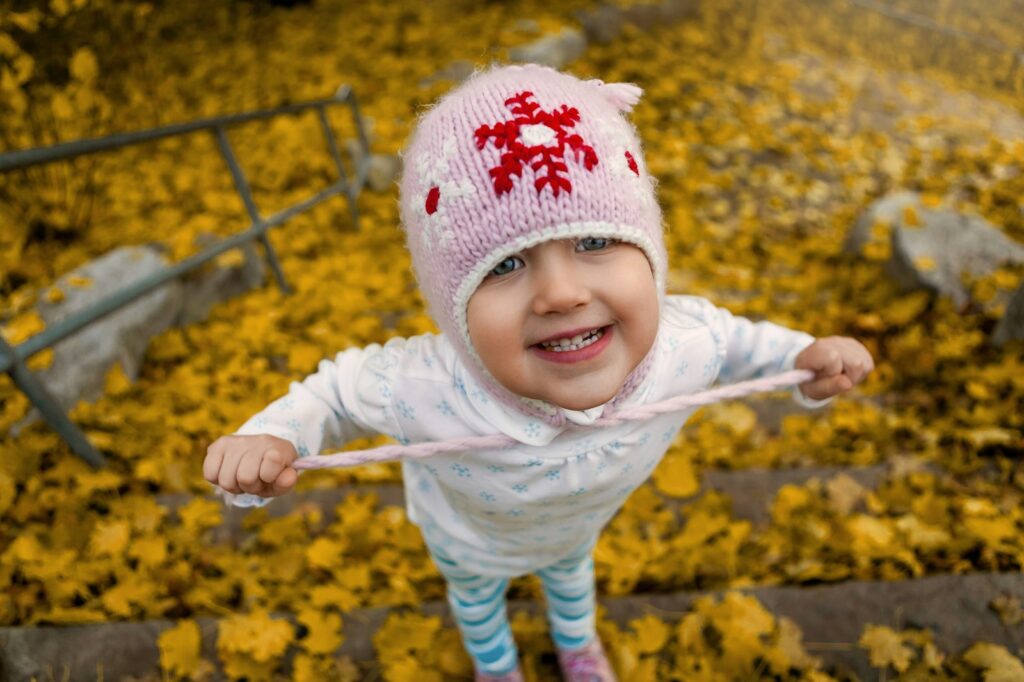 A smiling toddler in a pink knit hat stands among yellow autumn leaves.